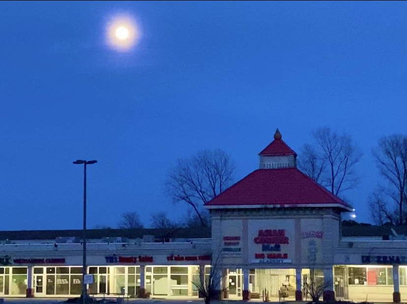 A strip mall with a decorated roof resembling asian architecture. There is a full moon above it, somber blue sky behind it.