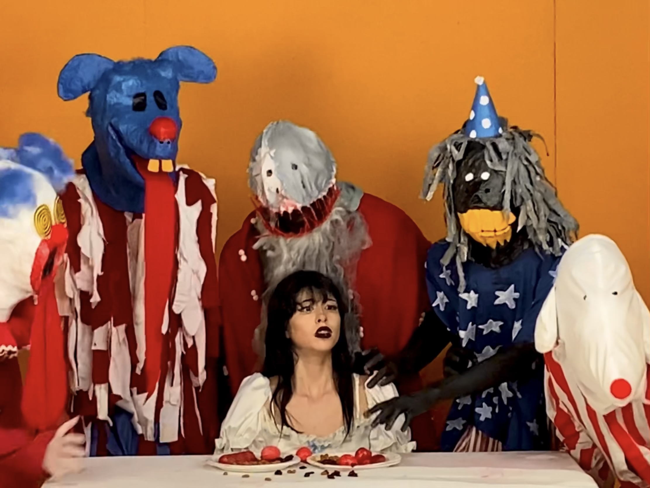 A sitting woman is surrounded by clowns at a table with food