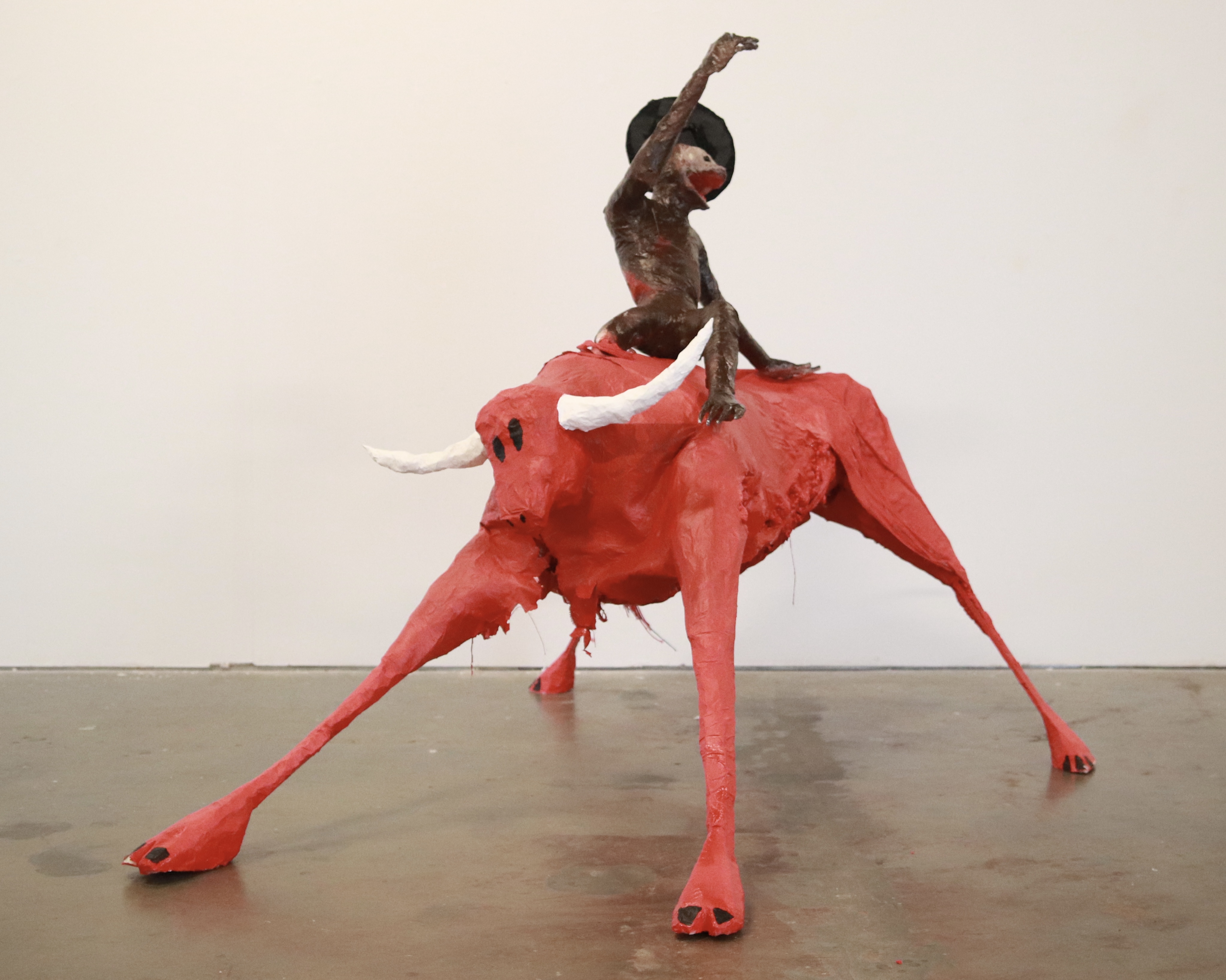 A small frightened monkey with a hat and a missing leg sits on a scared red bull